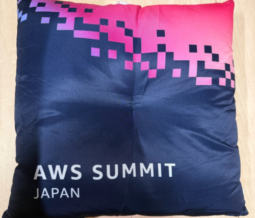 AWS Summit Japan 2026の基調講演早期入場特典として配布されたクッション。ネイビーの生地に「AWS SUMMIT JAPAN」の白文字ロゴが入り、角にピンクとパープルのピクセル柄があしらわれたデザイン。